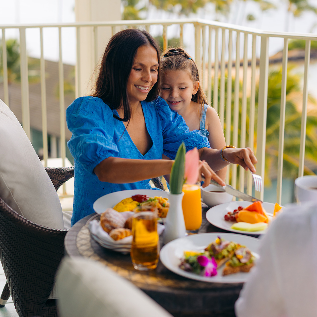 A woman and a young girl enjoy a meal on a balcony overlooking a tropical view.