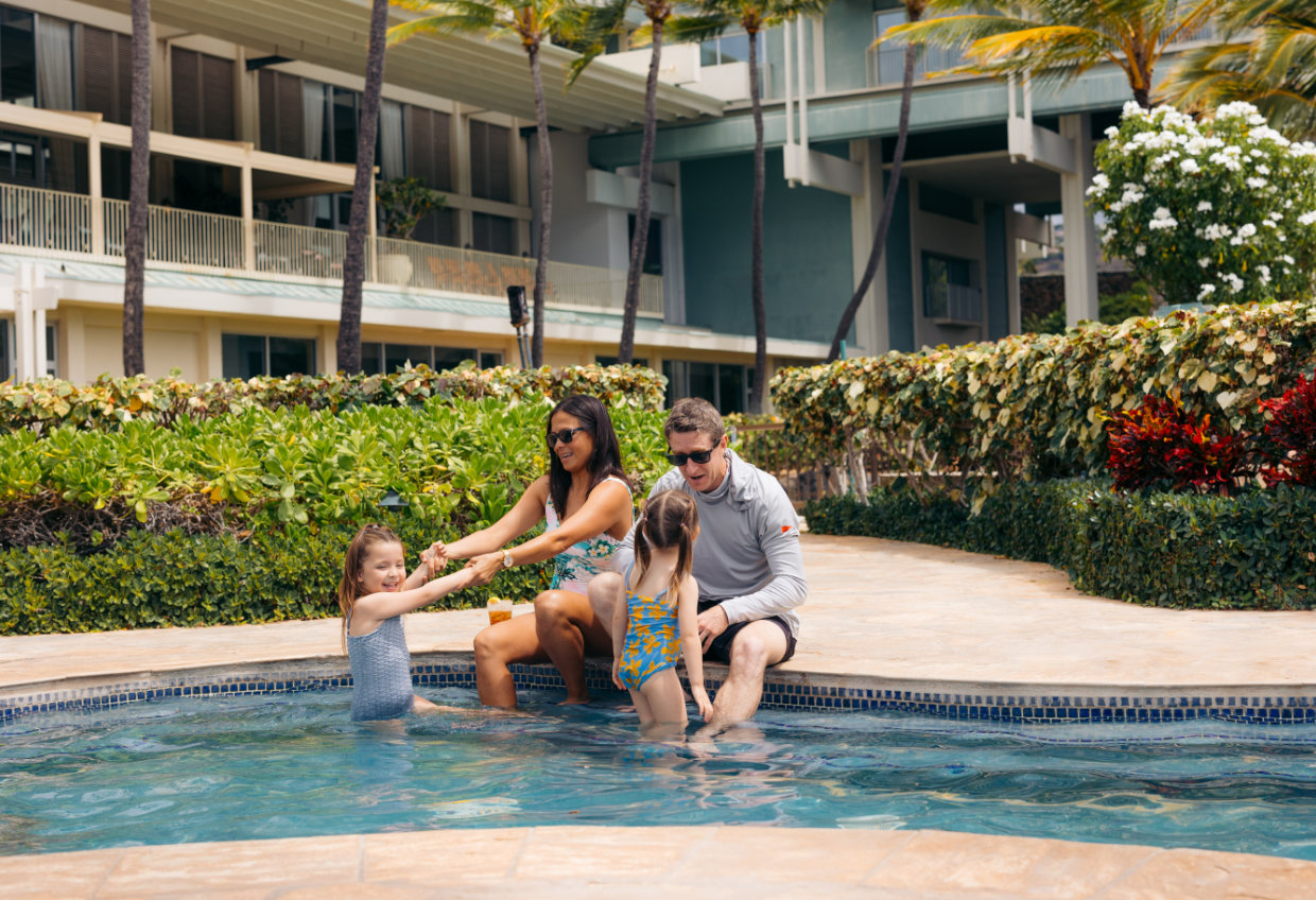 A family of four enjoys a moment together in a swimming pool with a hotel building in the background.