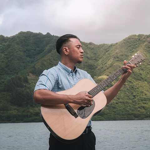 man holding guitar standing next to ocean with a mountain in the background